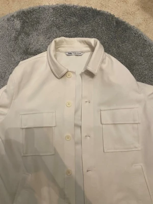 Vit overshirt från Zara - Stilren vit overshirt från Zara med klassisk krage och fyra knappar framtill. Två stora bröstfickor med lock och raka ärmar. Materialet är kraftigt och ger en snygg, strukturerad look. Perfekt att bära över en t-shirt för en avslappnad stil.