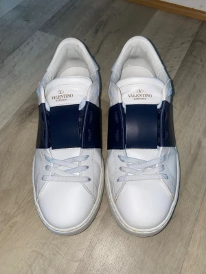 Valentino Garavani vita sneakers äkta - Snygga vita sneakers från Valentino Garavani med en bred mörkblå rem över mitten. Skorna är i läder och har ikoniska nitar på hälen. Klassisk låg modell med vita snören och diskret branding på plösen. Den är äkta såklart 