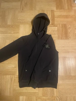 Svart Stone Island jacka - Säljer en svart jacka från Stone Island med dragkedja framtill och klassisk logotyp-patch på vänster ärm. Hoodien har två fickor med knappstängning och en luva. Passar XS-164.