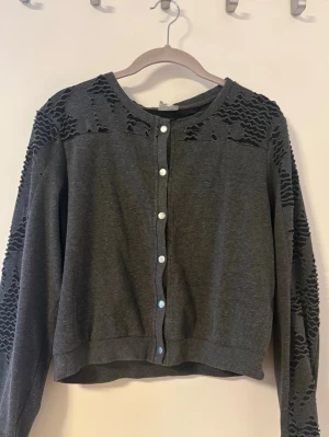 Mörkgrå cardigan med spetsdetaljer - Snygg mörkgrå cardigan med vita knappar framtill och coola spetsdetaljer på axlar och ärmar. Tröjan har rund halsringning och ribbade muddar vid ärmslut och nederkant. Perfekt för dig som gillar en unik twist på klassiska plagg.