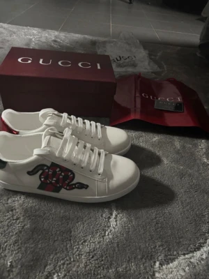 Gucci vita sneakers - Snygga vita sneakers från Gucci med klassiska gröna och röda ränder på sidan och ett coolt ormtryck. Skorna har lågt skaft, vita snören och är tillverkade i läder. Perfekta för dig som vill sticka ut med exklusiv streetstyle. Dem är helt nya o väntar på en ny ägare, storlek 42, för mer info skriv. 