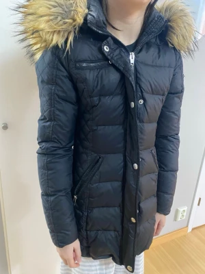 Svart lång dunjacka med fuskpäls - Snygg svart lång dunjacka med dragkedja och knappar framtill. Jackan har två snedställda fickor med dragkedja och en bröstficka. Stor huva med fluffig fuskpäls i brunt. Perfekt för kalla vinterdagar. Det finns ett hål på en armen som det synd på sista bilden. 