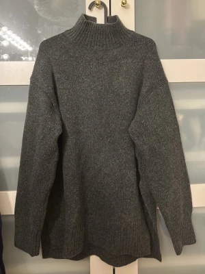 Mörkgrå stickad polotröja H&M - Mysig mörkgrå stickad polotröja från H&M med hög krage och avslappnad passform. Tröjan har långa ärmar och ribbade muddar vid hals, ärmslut och nederkant. Perfekt för kalla dagar och enkel att matcha med olika outfits.