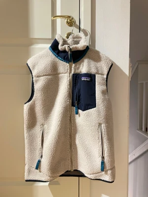 Beige fleeceväst från Patagonia - Mysig beige fleeceväst från Patagonia med marinblå detaljer. Västen har hög krage, dragkedja framtill, två sidofickor och en bröstficka med dragkedja. Perfekt för lager-på-lager och outdoor-vibe.