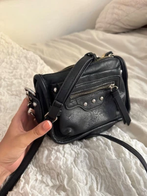 Svart axelväska med nitar och dragkedja - Snygg svart axelväska i skinnimitation med silverfärgade nitar och dragkedjedetaljer. Väskan har justerbar axelrem och ett mindre ytterfack framtill. Perfekt storlek för det viktigaste och ger en cool vibe till din outfit.❤️