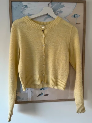 Gul kofta - Söt gul kofta med rund halsringning och pärlemorskimrande knappar framtill. Modellen är kortare med ribbade muddar vid ärmslut och nederkant. Perfekt att slänga över en topp för en fräsch look. Materialet känns mjukt och mysigt.