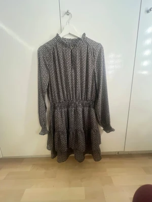 Svartvit mönstrad volangklänning - Kort klänning med svartvitt småmönster, volangdetaljer i kjolen och långa ärmar med lätt rynk vid ärmslut. Hög krage med liten volangkant och markerad midja. Perfekt för dig som gillar en söt och trendig stil.