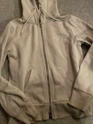 Beige hoodie från GANT med dragkedja - Snygg beige hoodie från GANT med dragkedja framtill och broderad logga på bröstet. Hoodien har huva med dragsko, ribbade muddar och två fickor fram. Tillverkad i mjukt material som känns skönt mot huden. Perfekt för en avslappnad och stilren look.