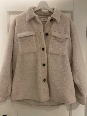 Beige teddyjacka med fickor - Mysig beige teddyjacka med stora bruna knappar och två rejäla bröstfickor med lock. Jackan har en avslappnad passform, klassisk krage och är gjord i fluffigt teddy-material som håller dig varm. Perfekt för dig som gillar en trendig och bekväm stil. Strl 176  ca16 år ungefär som XS