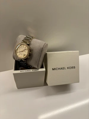 Guldig klocka från Michael Kors - Snygg guldig klocka från Michael Kors med länkarmband och glittrande stenar runt urtavlan. Klockan har flera små urtavlor och datumvisning. Kommer i originalask med logga. Perfekt accessoar för dig som gillar stilrena detaljer.