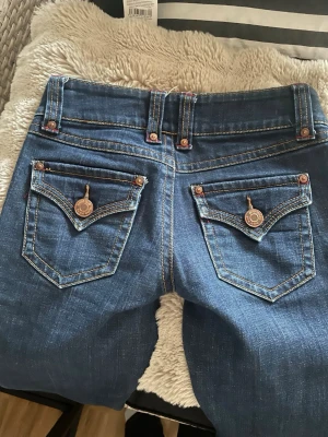 Blå Gina Tricot jeans med detaljer - Snygga blå jeans från Gina Tricot med dekorativa ficklock och metallknappar bak. Klassisk femficksmodell med kontrastsömmar och låg midja. Jeansen har coola detaljer som hängande metallbrickor på bakfickorna och röda sömmar vid fickorna fram.