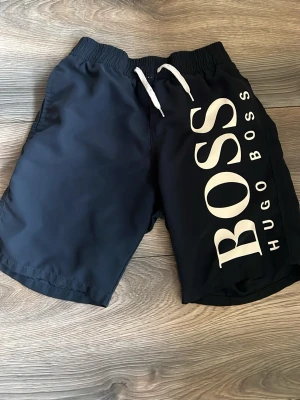 Marinblå badbyxor från Hugo Boss - Snygga marinblå badbyxor från Hugo Boss med stort vitt BOSS-tryck på ena benet och vita dragsnören i midjan. Klassisk modell med elastisk midja och innerbyxa i mesh. Perfekta för stranden eller poolen.