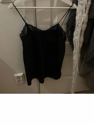 Svart spetslinnetopp med tunna band - Snygg svart linnetopp med tunna axelband och spetsdetaljer upptill. Perfekt för att styla lager på lager eller bära som den är. Tidlös och enkel design som passar till mycket.