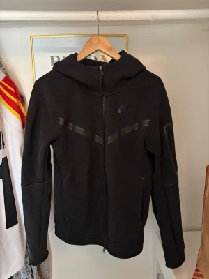 Svart Nike hoodie med dragkedja - Svart hoodie från Nike med hel dragkedja och huva. Snygga detaljer med svarta band på bröstet och en ficka med dragkedja på ärmen. Perfekt för dig som gillar sportig och stilren design. Materialet är mjukt och bekvämt.