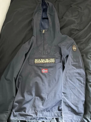 Mörkblå vindjacka från Napapijri - Snygg mörkblå vindjacka från Napapijri med huva och halv dragkedja framtill. Stor ficka på bröstet med Napapijri-logga och norsk flagga. Patch med logga på ärmen. Jackan är tillverkad i slitstarkt material och har en sportig, avslappnad passform.