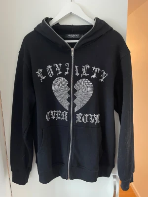 Svart hoodie från Trotherz  - As snygg och unik Svart zip-hoodie från trotherz med  huva och stort tryck med texten 'Loyalty Over Love' och ett trasigt hjärta i pärlor! Har en mjuk och skön bomullskänsla, två fickor fram och dragkedja hela vägen upp. Hoodien är använd fåtal gånger i väldigt bra kvalite med alla pärlorna kvar på plats!