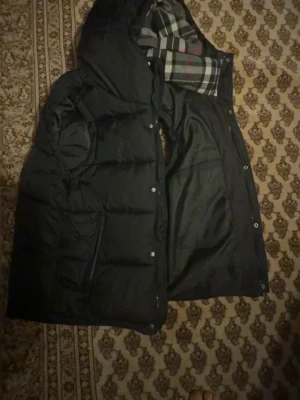 dunjacka - Snygg svart dunjacka med huva och rutigt foder i svart, vitt och rött. Jackan har dragkedja och tryckknappar framtill samt praktiska fickor. Perfekt för kalla vinterdagar och har en klassisk, tidlös look. Storlek: L men passar som M.