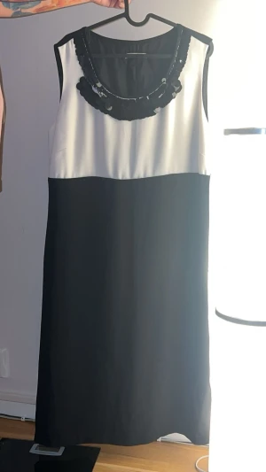 Svartvit ärmlös klänning - Snygg svartvit klänning med ärmlös design och rak passform. Överdelen är vit med en dekorativ svart detalj vid halsen, medan nederdelen är helsvart. Perfekt för dig som gillar stilrena kontraster och enkel elegans.