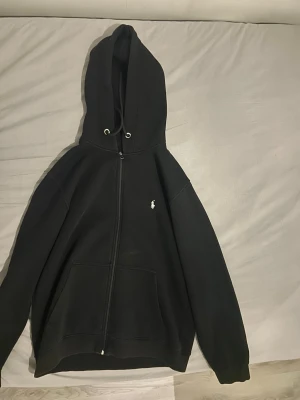 Svart zip hoodie från Polo Ralph Lauren scannen är borta - Svart hoodie med dragkedja från Polo Ralph Lauren. Klassisk design med huva och vita dragsnören, samt den ikoniska broderade loggan på bröstet. Stora fickor framtill och mjukt material som känns skönt mot huden. Perfekt för en avslappnad och stilren look.