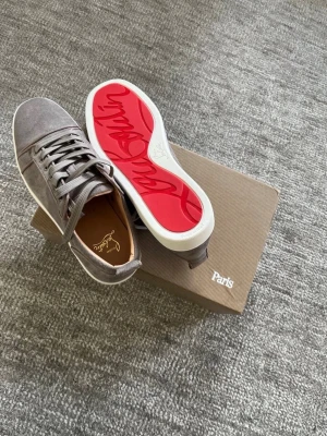 Gråa Loubs från Christian Louboutin - Snygga gråa sneakers från Christian Louboutin i mocka med den ikoniska röda sulan och vita kanter. Skorna har klassisk snörning och diskret logga på insidan. Perfekta för dig som vill ha en exklusiv och trendig look. Helt nya, oanvända.