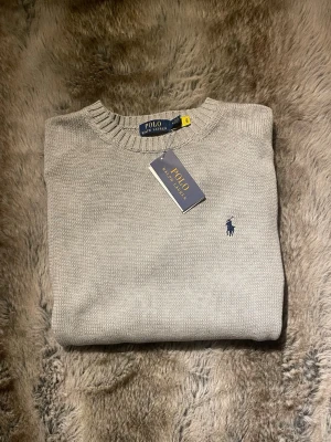 Ljusgrå stickad tröja Polo Ralph Lauren - Ljusgrå stickad tröja från Polo Ralph Lauren med rund halsringning och klassisk blå broderad logga på bröstet. Tröjan har ribbade muddar vid hals, ärmslut och nederkant. Tillverkad i mjukt material för en clean och stilren look.
