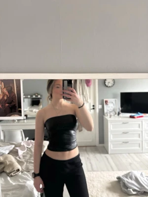 Svart axelbandslös tubtopp i skinnimitation - Snygg svart axelbandslös tubtopp i skinnimitation. Toppen är croppad och har en tight passform som sitter snyggt mot kroppen. Perfekt för dig som vill ha en edgy och trendig look.