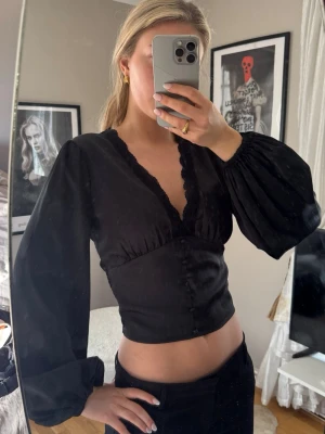 Svart satin topp puffärmar och spets - Snygg svart topp med djup v-ringning och dekorativ spetskant. Modellen har puffiga långa ärmar och smock baktill för en bekväm passform. 