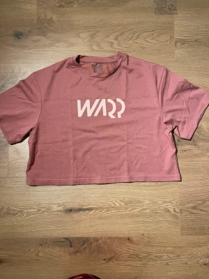 Rosa croppad t-shirt från Warp - Säljer en rosa croppad t-shirt från Warp med vit logga framtill. T-shirten har rund halsringning och korta ärmar. Tillverkad i mjuk bomull som känns skön mot huden. Perfekt för en avslappnad och trendig look.