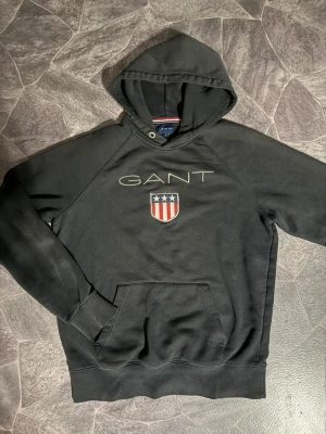 GANT hoodie  - Svart hoodie från GANT med stor broderad logga och amerikanskinspirerat sköldmärke på bröstet. Klassisk känguruficka framtill och justerbar huva med snörning. Tillverkad i mjuk bomullsblandning för skön känsla och avslappnad passform.
