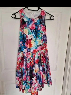 Färgglad blommig klänning från Ginger Fizz - Superfin ärmlös klänning med färgstarkt blommönster i rosa, blått, rött och vitt. Modellen är kort med rund halsringning och intressant rygg. Perfekt för sommarens alla tillfällen!