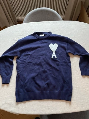 Mörkblå stickad tröja Ami Paris - Mörkblå stickad tröja från Ami Paris med vitt hjärta och bokstaven A på bröstet. Tröjan har rund halsringning, ribbade muddar och nederkant. Perfekt för dig som gillar stilrena detaljer och vill ha något bekvämt och snyggt.