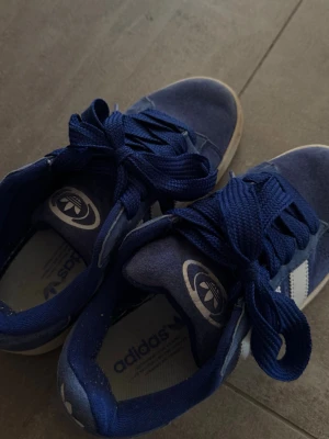 Blå Adidas Campus sneakers - Säljer ett par blå Adidas Campus sneakers med klassiska vita ränder på sidorna och Adidas-logga på plösen. Skorna har en låg siluett, breda blå skosnören och ovandel i mocka. Inte använda mycket alls, 3 gånger.