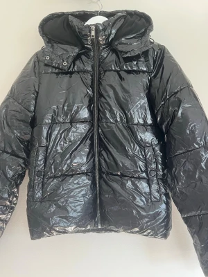Svart glansig pufferjacka ifrån Nelly  - Säljer denna svarta glansiga pufferjackan med huva samt foder inuti ifrån Nelly i storlek xxs. Använd ett fåtal ggr men inget som syns!