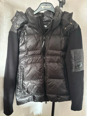 C.P Company hybrid jacka  - Säljer en svart dunjacka från C.P. Company med glansig finish, stickade ärmar och ikonisk linsdetalj på ena ärmen. Jackan har huva, dragkedja framtill och flera fickor. Jackan är i stolen XS men skulle även passa de som har S, tags osv medföljer