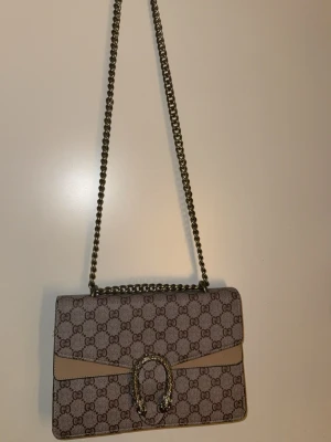  axelväska beige - Snygg  Dionysus axelväska i beige och brunt med klassiskt GG-mönster. Väskan har en kedjerem i metall, dekorativt spänne framtill och flera fack inuti. Tillverkad i canvas och läder med exklusiva detaljer.