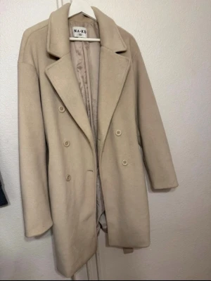Beige kappa från NA-KD  - Stilren beige kappa från NA-KD med dubbelknäppning och klassisk krage. Använd men välvårdad och i fint använt skick. Storlek 36. Nypris 1800 kr. 