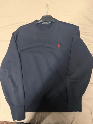 Mörkblå sweatshirt från Polo Ralph Lauren - Mörkblå sweatshirt från Polo Ralph Lauren med klassisk rund halsringning och röd broderad logga på bröstet. Tröjan har ribbade muddar vid ärmslut och nederkant. Perfekt för en avslappnad och stilren look.