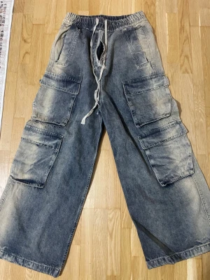 Baggy cargobyxor i denim med snörning - Säljer ett par baggy cargobyxor i tvättad blå denim med bred passform och elastisk midja med snörning. Byxorna har flera stora cargofickor på sidorna och dragkedja fram. Perfekta för en avslappnad streetwear-look.