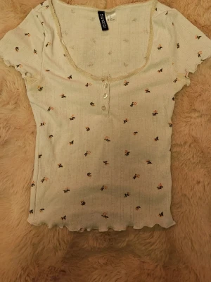 Blommig vit topp  - Supersöt vit topp från h&m med små blommor, korta ärmar och dekorativa knappar framtill. Toppen har en söt spetskant vid halsen och vågig kant nedtill. Perfekt för dig som gillar lite downtown girl stil ☕🎀📚