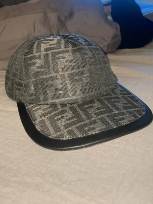  Fendi keps - Säljer en grå keps från Fendi med det klassiska FF-logomönstret över hela kepsen. De enda tråkiga är att bokstaven (e) är borta från ”Fendi” Ibak annars inga slitningar.