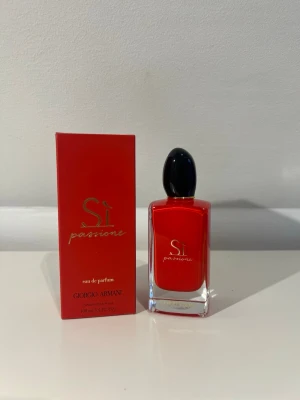 Sì Passione parfym från Armani - Sì Passione från Giorgio Armani är en elegant parfym i en snygg röd glasflaska med svart kork. Flaskan har en modern, rektangulär form och guldtext på framsidan. Förpackningen är också röd med guldiga detaljer, vilket ger en lyxig känsla.