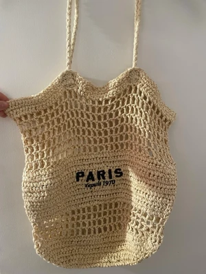 Beige virkad strandväska Paris - Snygg beige strandväska i virkad stil med texten 'PARIS depuis 1970' framtill. Väskan har ett nätmönster och långa, tvinnade axelband. Perfekt för att bära med sig till stan eller stranden. Materialet känns naturligt och luftigt. Snyggväska till stranden. 