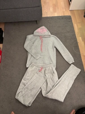 Synaworld set - Säljer ett par grå mjukisbyxor med resår i midjan och rosa dragsko. Tillhörande hoodie med rosa tryck på huvan och matchande rosa snören. Perfekt för chill dagar hemma eller när du vill vara extra bekväm.
