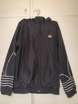 Svart vindjacka från Adidas - Snygg svart vindjacka från Adidas med vit logga på bröstet och vita grafiska detaljer på ärmarna. Jackan har dragkedja framtill, huva och elastiska muddar vid ärmar och midja. Perfekt för en sportig och avslappnad stil.