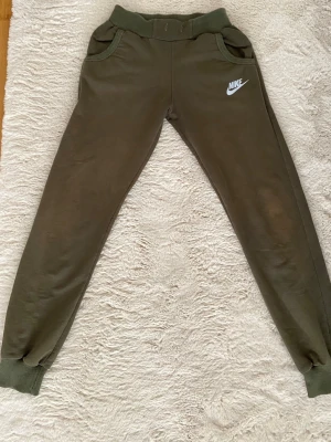 Olivgröna mjukisbyxor från Nike dri-fit - Säljer ett par olivgröna mjukisbyxor från Nike med resår i midjan och muddar vid bensluten. Byxorna har två fickor fram och en vit Nike-logga på vänster lår. Perfekta för chill dagar eller träning.