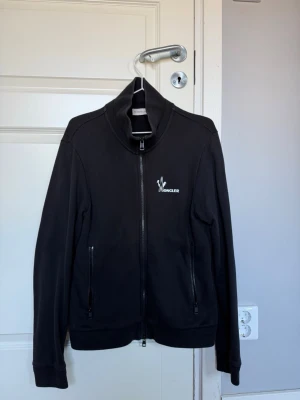 Svart zip hoodie från Moncler - Säljer en svart hoodie från Moncler med hel dragkedja framtill och två fickor med dragkedja. Tröjan har hög krage och Moncler-logga i vitt på bröstet. Tillverkad i mjukt bomullsmaterial som känns skönt mot huden.