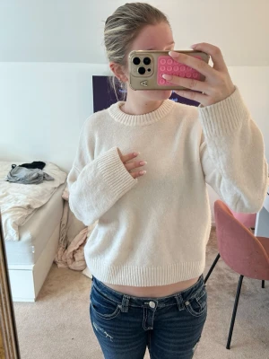 Krämvit stickad tröja med rund hals - En enkel och stilren krämvit stickad tröja med rund hals och ribbade muddar. Tröjan har en något boxig passform och långa ärmar, perfekt för lager-på-lager. Materialet ser ut att vara mjukt och bekvämt, idealiskt för kyliga dagar.