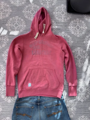  hoodie från Superdry - Säljer en hoodie från Superdry med tryck på bröstet och klassisk känguruficka. Hoodien har vita dragsnören och ribbade muddar vid ärmar och nederkant. Perfekt för en avslappnad och cool stil. Materialet är mjuk bomullsblandning.