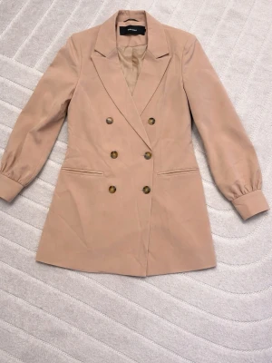 Beige dubbelknäppt kavaj - Stilren beige kavaj från veromoda med dubbelknäppning och fyra bruna knappar framtill. Kavajen har markerade axlar, långa ärmar med lätt puff och två fickor framtill. Materialet är slätt och ger en elegant look. Perfekt för att lyfta din outfit.