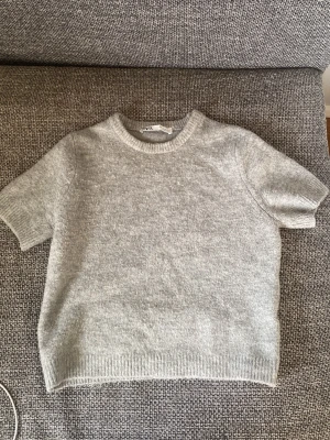 Grå stickad kortärmad tröja från Zara  - Mysig grå stickad tröja från Zara  med korta ärmar och rund halsringning. Tröjan har en enkel och stilren design med ribbade kanter vid ärmslut och nederkant. Perfekt för lager-på-lager under kyliga dagar.